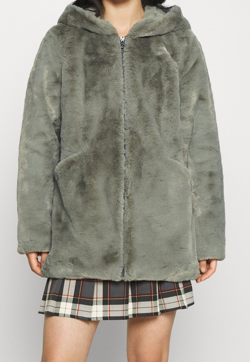 En person som bär en grå huva med fuskpälsjacka som är dragkedja, ihop med en plisserad kjol i svart, vitt och rött rutmönster som är synlig nedan.