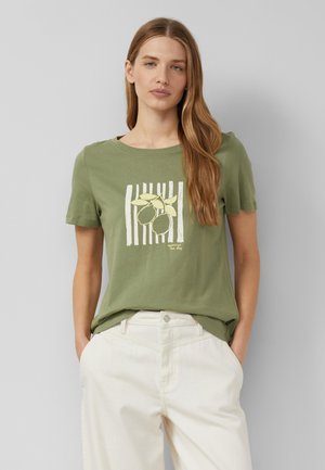 Femme aux longs cheveux châtain clair porte un t-shirt vert olive avec un motif citron et un pantalon blanc, debout devant un fond gris clair uni.