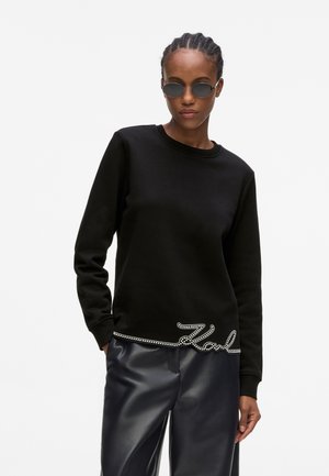 Persoon die een zwarte sweater met ronde hals draagt met een "Karl" strass-steendetail aan de zoom, zwarte leren broek en ronde donkere zonnebril.