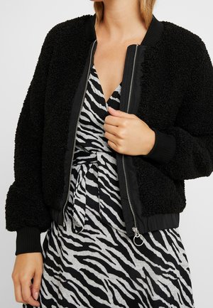 Persona che indossa una giacca nera testurizzata con zip sopra un vestito a stampa zebra bianco e nero con vita annodata.