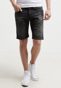 Hombre que lleva pantalones cortos de mezclilla negros desgastados, una camiseta blanca y zapatillas altas blancas, de pie con una mano en el bolsillo.