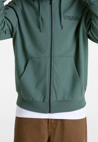 Sudadera con capucha verde con cremallera, capucha forrada, puños acanalados y bolsillos frontales. Presenta un logotipo bordado de "Vans" en verde a juego.