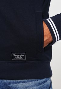 Felpa navy con colletto e polsini a coste, con strisce bianche, tasca a marsupio e etichetta cucita all'orlo. Tessuto di cotone.