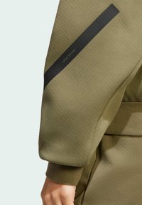 Olivovo zelený športový top s textúrovanou tkaninou, s čiernym akcentovým pruhom pozdĺž rukáva a rebrovanými manžetami.