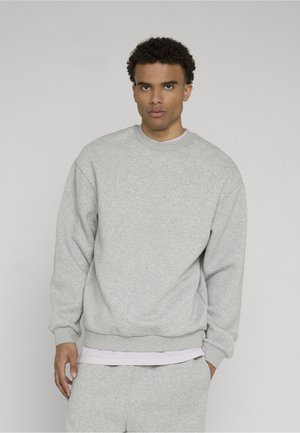 Ung mand med kort krøllet hår iført en lysegrå sweatshirt og matchende joggingbukser foran en ensfarvet baggrund.