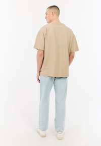 Beige katoenen t-shirt met korte mouwen, recht model. Draag met lichtblauwe ontspannen fit jeans en witte sneakers. Effen achtergrond.