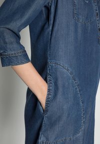 Tummansinisessä farkkupaidassa on nappirivistö edessä, rintatasku, käännetyt hihat ja sivutaskut, joissa on kontrastivärinen ommel.