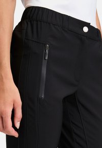 Czarne spodnie z elastycznego materiału z elastycznym paskiem w talii, boczną kieszenią na zamek oraz przeszyciami wzdłuż boków.