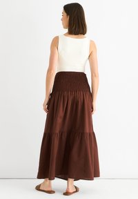 Brauner Maxikleid mit gestuftem Design, gerafftem Bund und fließendem Stoff. Getragen mit einem enganliegenden weißen Oberteil und braunen Sandalen, von hinten gezeigt.