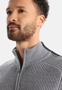 Grauer Zip-Pullover mit strukturiertem Rippdesign, hohem Kragen und schwarzen Akzenten an den Schultern. Der Stoff wirkt weich und formstabil.
