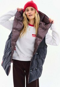 Žena v červenej čiapke, bielej mikine s červeným lemovaním, tmavej nadýchanej vesty a tmavých nohaviciach, ktorá si upravuje golier vesty na bielom pozadí.