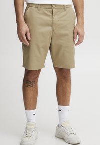 Khaki-Shorts aus Baumwolle, gerade geschnitten, mit Fronttaschen, Knopfverschluss, knielang, kombiniert mit weißen Turnschuhen und Socken.