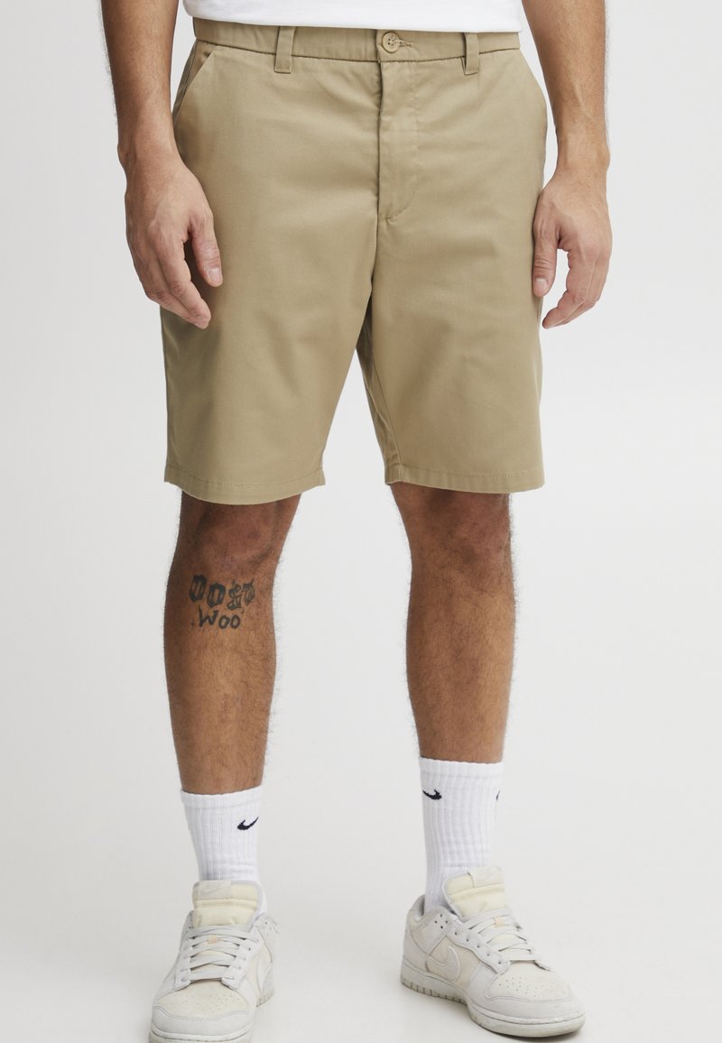 Khaki-Shorts aus Baumwolle, gerade geschnitten, mit Fronttaschen, Knopfverschluss, knielang, kombiniert mit weißen Turnschuhen und Socken.