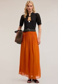 Svart topp med texturerade ärmar, kombinerad med en flödig orange maxikjol. Tillbehör inkluderar ett svart bälte och en vävd handväska.