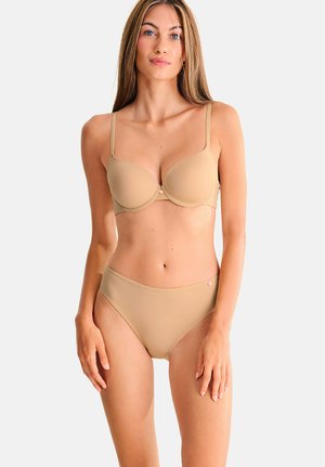 Femme aux longs cheveux blonds portant un soutien-gorge beige et une culotte taille haute assortie, debout devant un fond clair uni.