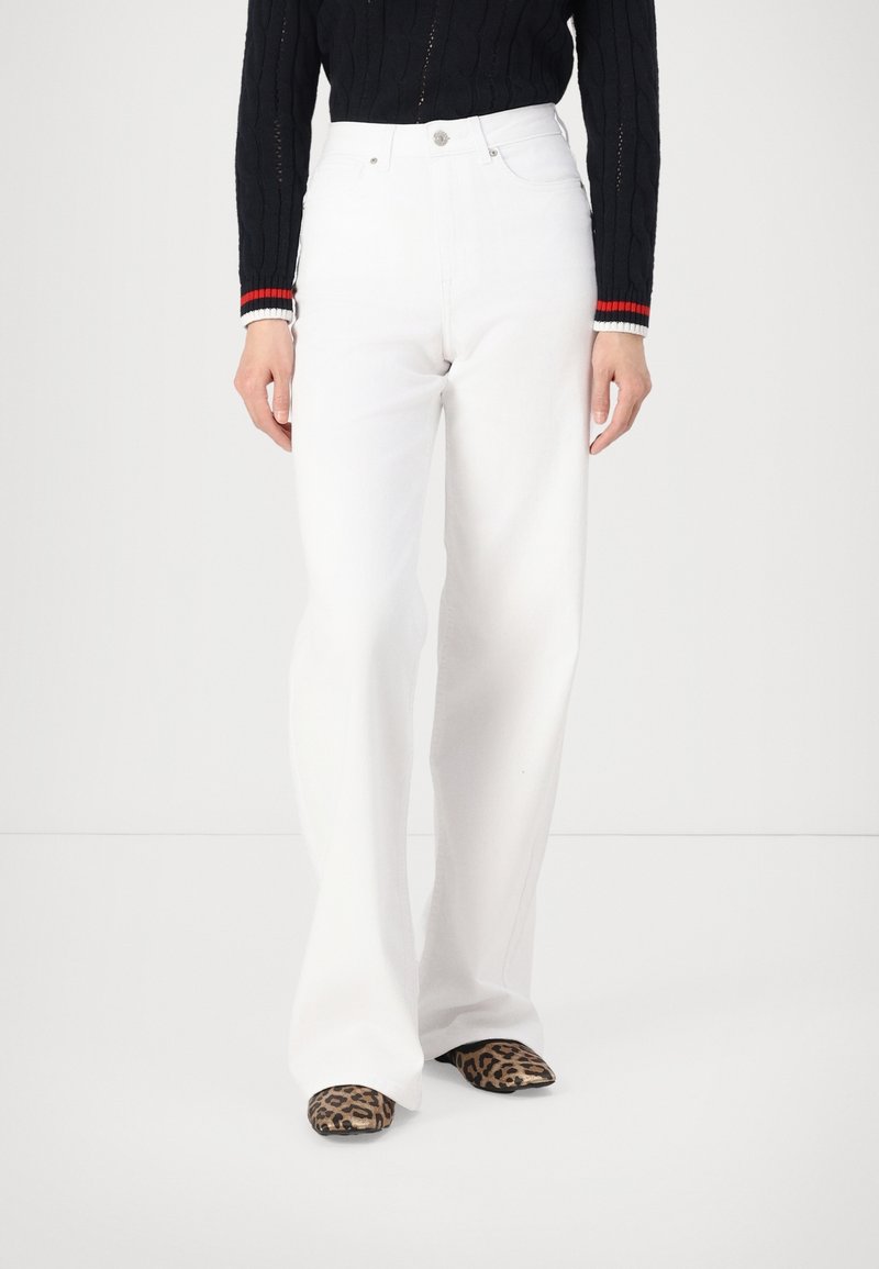 Personne portant un pantalon blanc taille haute à jambes droites, un pull noir avec des poignets à rayures rouges et blanches, et des chaussures imprimé léopard, debout.