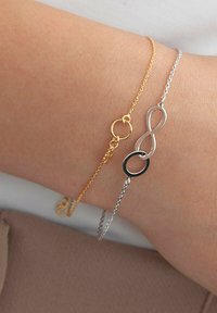 Złote i srebrne bransoletki łańcuszkowe na nadgarstku; jedna z nich ma symbol nieskończoności oraz czarny okrągły akcent, a druga jest prostym okrągłym ogniwem.