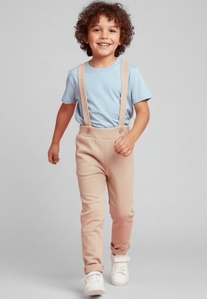 Jasnoniebieska koszulka, beżowe dopasowane spodnie na szelkach i białe sneakersy. Spodnie mają dwa guziki i gładką teksturę.