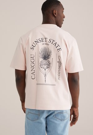 Mann trägt ein hellrosa T-Shirt mit schwarzem botanischem Druck und den Texten "Sunset State of Mind", "Canggu" und "Afternoons" auf der Rückseite.
