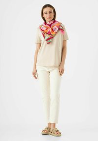 Beige T-shirt, witte jeans en geweven sandalen. Het model draagt een kleurrijke bloemen sjaal met rozee, oranje en groene accenten.