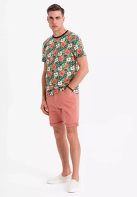 Blumen T-Shirt mit kurzen Ärmeln auf grünem Hintergrund, mit roten und weißen Blumen. Kombiniert mit rosa Shorts und weißen Schlupfschuhen.