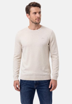 Homme portant un pull à manches longues beige clair et un jean bleu, regardant légèrement sur le côté devant un fond blanc uni.