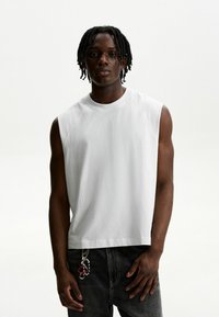 Hvid ærmeløs T-shirt med rund hals, der har blødt stof og en afslappet pasform. Båret med mørke jeans og en nøgleringsaccessory.