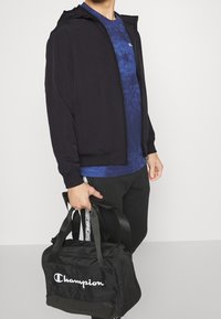 Black zip-up jacket, blue marbled t-shirt, black pants, holding a black Champion duffel bag with white logo.