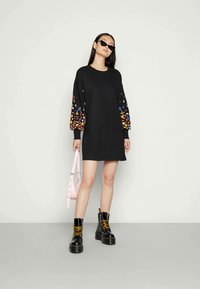 Donna che indossa un mini abito nero con maniche a fiori colorati, stivali platform neri con lacci gialli, occhiali da sole a occhio di gatto e tiene una borsa rosa.