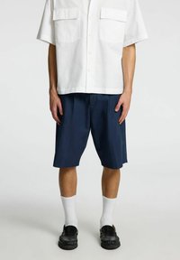 Hvid kortærmet skjorte med to forreste lommer, parret med marineblå shorts. Modellen bærer hvide sokker og sorte loafers med tekstureret detalje.