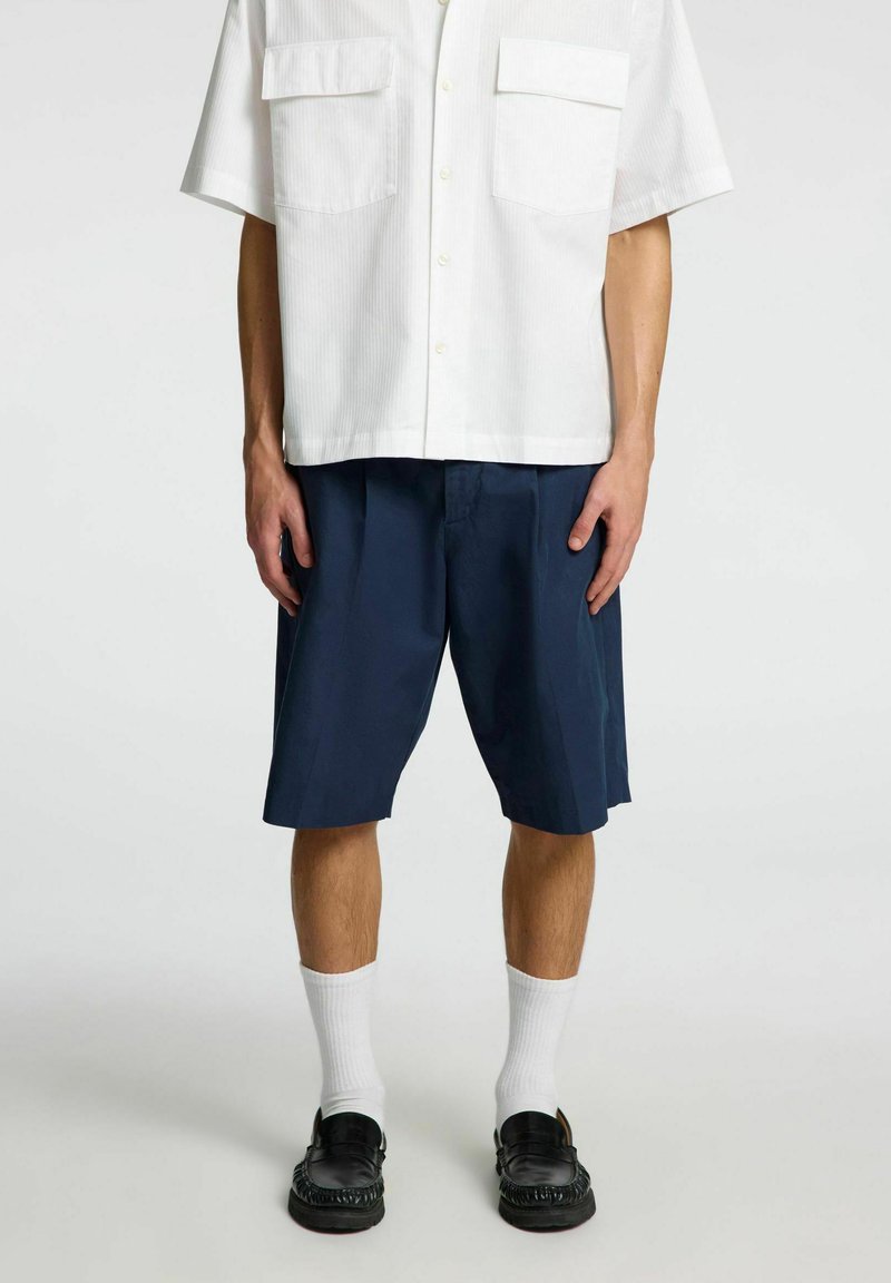 Hvid kortærmet skjorte med to forreste lommer, parret med marineblå shorts. Modellen bærer hvide sokker og sorte loafers med tekstureret detalje.