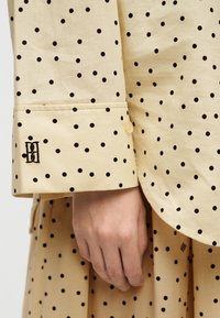Main reposant près du poignet d'une chemise beige à pois noirs avec un logo noir brodé sur le poignet.
