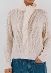 Cardigan lavorato a maglia beige con motivo di punto aperto, polsini a coste e dettaglio della sciarpa legata al collo. Dotato di chiusura con bottoni e vestibilità rilassata.