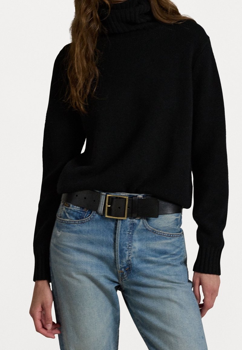 Femme portant un pull à col roulé noir rentré dans un jean bleu clair avec une ceinture noire avec une boucle carrée dorée.