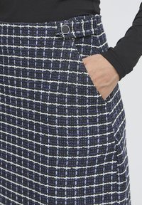 Mână în buzunarul fustei din tartan albastru cu talie înaltă, cu un brâu butonat negru, purtată cu un top negru cu mânecă lungă.