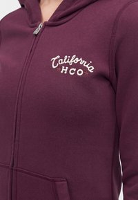Maroon zip-up hoodie with white "California HCO" embroidery on the chest and a front pocket visible at the bottom.