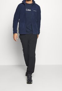 Marinblå fleecejacka med hög krage och dragkedja, tillsammans med en marinblå t-shirt och svarta byxor, bärs med svarta träningsskor.