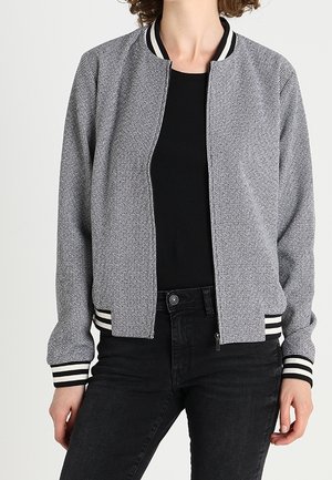 Vrouw draagt een grijze bomberjack met patroon, zwarte en witte gestreepte boorden en kraag, over een zwart shirt en zwarte jeans.