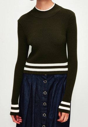 Mørkegrøn ribbet langærmet sweater med hvide striber på manchetter og talje, kombineret med en mørkeblå nederdel med knapper.
