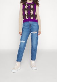 Stickad tröja i lila, brunt och crème med rutor, lager-på-lager över en beige polotröja, kombinerad med blå trasiga jeans med raka ben och beigea sneakers.