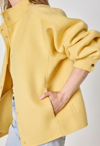 Yellow fleece jacket with a high collar, snap closures, and side pockets. Features wide sleeves and a soft texture with a relaxed fit.