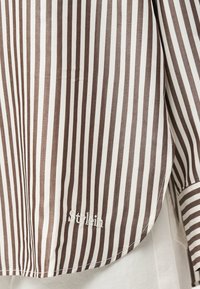 Καφέ και λευκή κάθετα ριγωτή λάμψη πουκάμισου με λευκό κεντημένο κείμενο με καλλιγραφικά γράμματα "Stylein" κοντά στο κάτω άκρο.