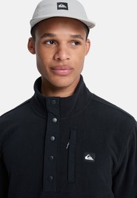 Sudadera de forro polar negra con cuello alto, botones a presión, bolsillo con cremallera y un pequeño parche de logo en blanco. Gorra de color claro con agujeros de ventilación.