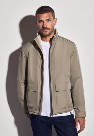 Mann mit kurzem Haar und Bart, der eine beige Zip-Jacke, ein weißes Hemd und dunkle Jeans trägt, steht vor einer hellen Wand mit Schatten.