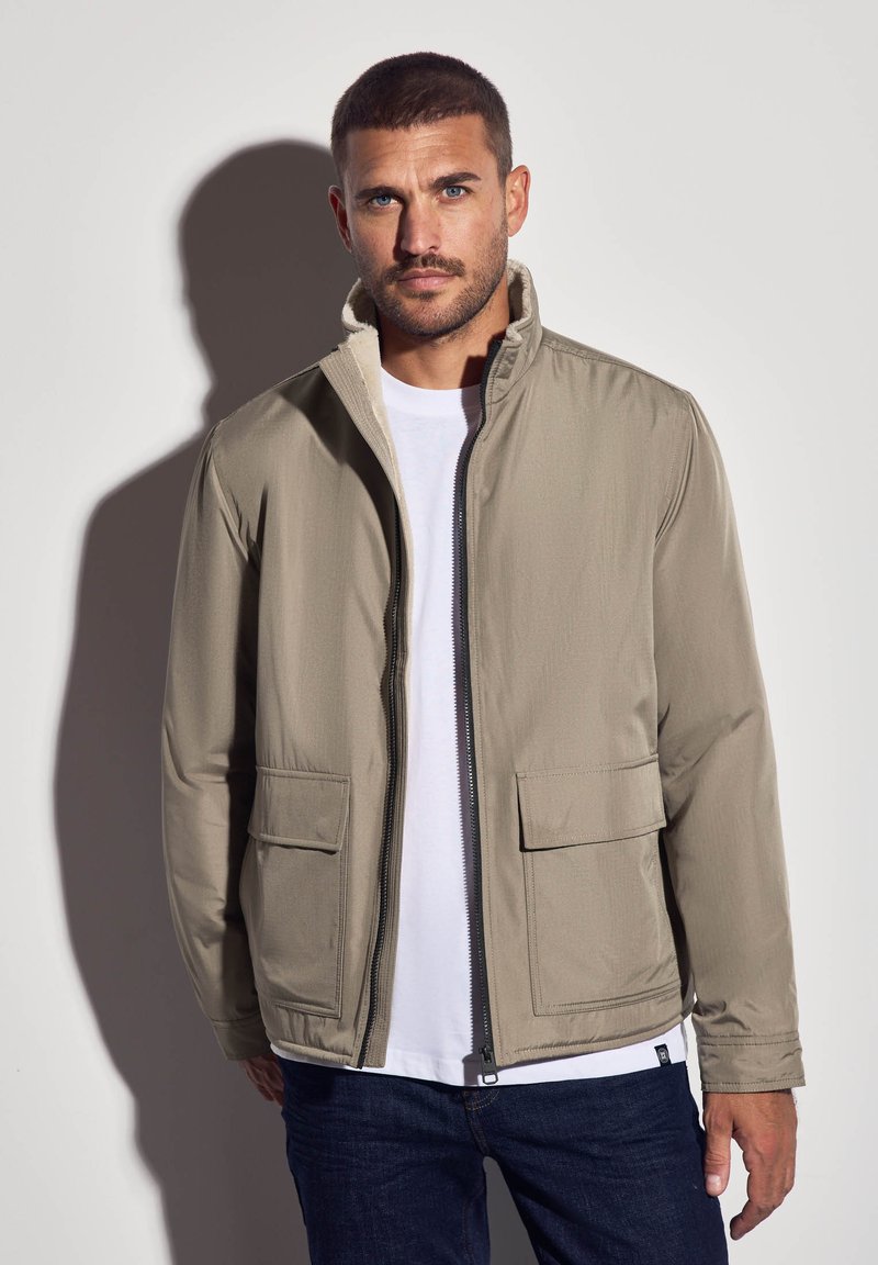 Homme aux cheveux courts et à la barbe, portant une veste zippée beige, une chemise blanche et un jean noir, se tenant contre un mur clair avec une ombre projetée.