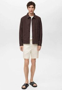 Brun knapjakke med hvid skjorte under, beige shorts og sorte sandaler. Enkel design med lommer på jakken.