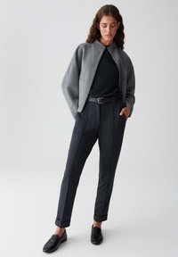 Woman standing in gray jacket, black top, dark tailored pants, black belt, and black loafers against a plain light background.