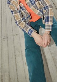 Camicia a quadri beige, blu e arancione su una felpa arancione brillante; pantaloni turchesi; mani unite, seduto su una superficie in legno testurizzata.