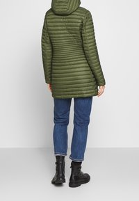 Doudoune vert olive avec capuche, présentant des coutures verticales et une coupe ajustée. Portée sur un jean bleu et des bottes noires.