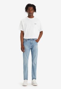 Mies seisoo yhdellä kädellä taskussa, päällä valkoinen Levi's t-paita, vaaleansiniset farkut ja valkoiset tennarit yksinkertaisella taustalla.